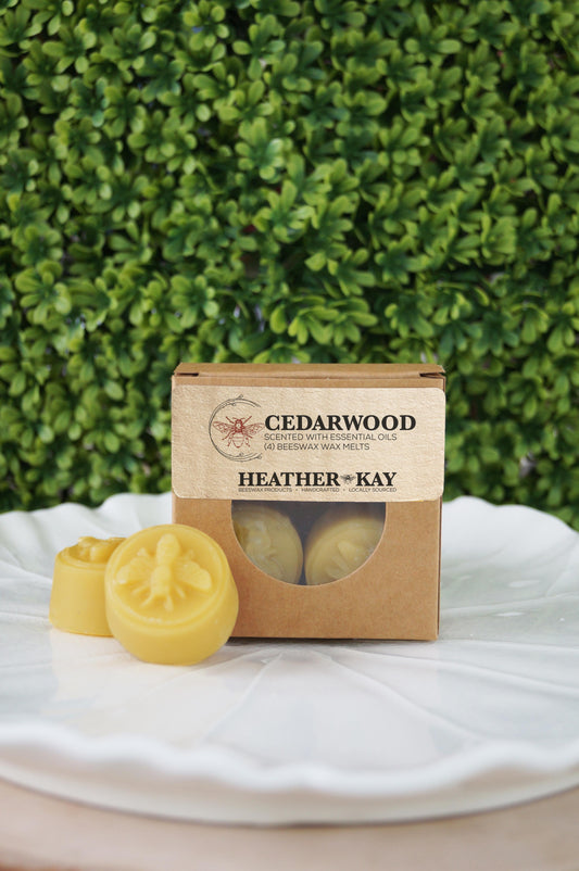 Box of handcrafted Cedarwood beeswax melts by Heather Kay Candles, with two yellow beeswax wax melts displayed on a white ceramic leaf dish in front of a green plant background.