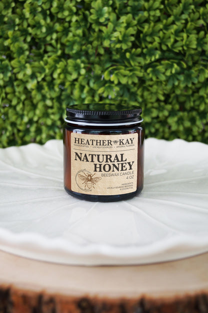 A black candle jar with a label reading 'Heather Kay Natural Honey' placed on a white surface with greenery in the background.